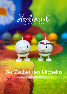 Ein weißer Hoptimist und der Mittsommer-Hoptimist mit Blumenkranz auf dem Kopf vor dem Juhannustaika-Stoff von Marimekko und vielen weiteren bunten Hoptimisten im Hintergrund. 
