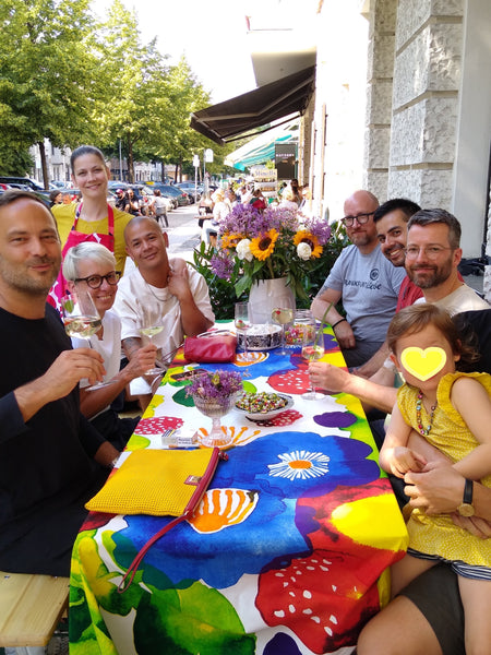 KundInnen und FreundInnen bei der Mittsommerfeier von HARTOG Berlin. Alle sitzen am geschmückten Tisch, der Stoff der Tischdecke heißt Juhannustaika von Marimekko. 