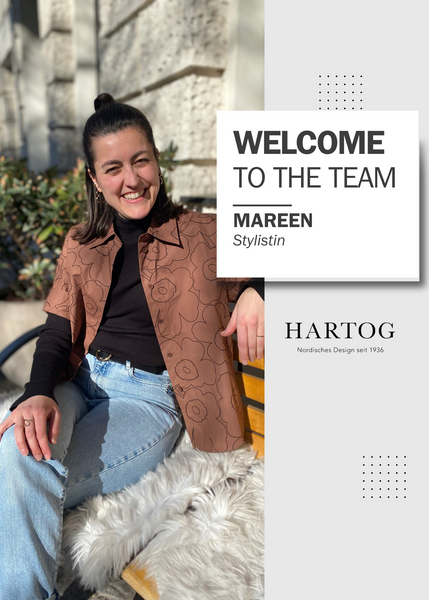 Neu im Team: Mareen. Hier sitzt sie auf der HARTOG-Bank und lächelt, trägt einen High Bun, eine helle Jeans, ein schwarzes Longsleeve und darüber eine kurzärmlige Piirto Unikko Bluse in braun von Marimekko.  