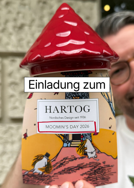 Unsere Einladung zur Moomin's Day Feier am 08.08.26: Torben hält die Karte und das Dach des Moominhauses darüber. Das Moominhaus ist auf der Moomin's Day Tasse 2026 zu sehen.